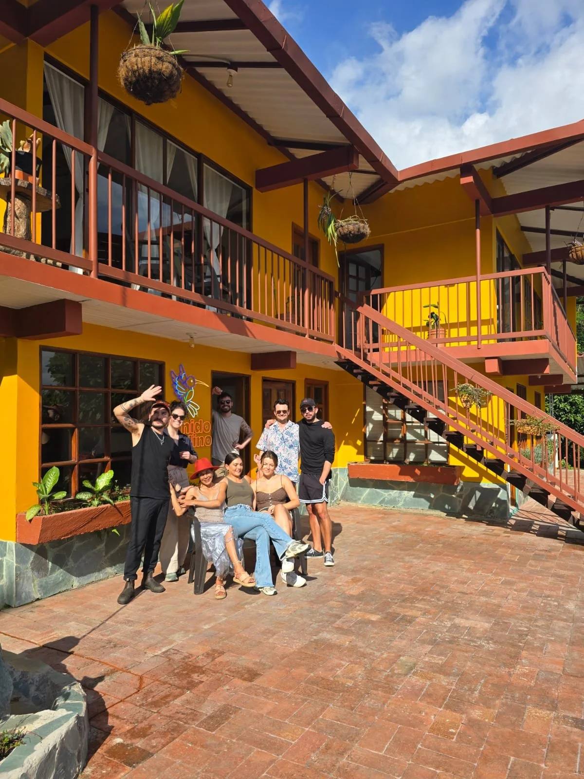 Grupo de siete personas posando frente a un edificio de color amarillo con balcones y escaleras exteriores. La escena es al aire libre con cielo despejado y nublado.