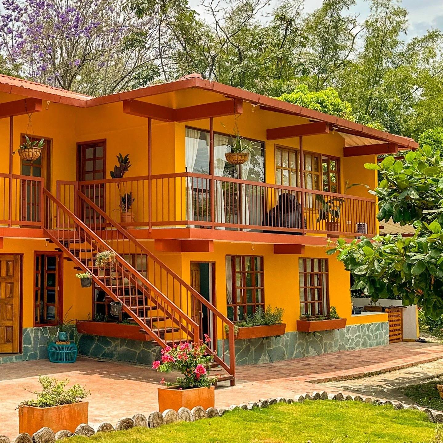 Casa amarilla con segundo piso y balcón, rodeada de árboles verdes y plantas