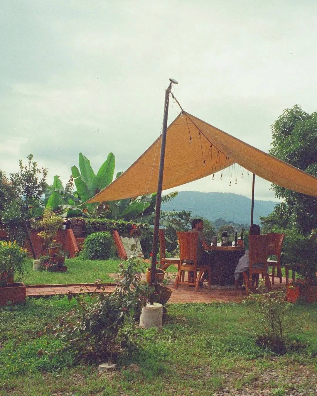 Personas sentadas en una mesa bajo una sombrilla en el jardín con plantas y vista a las montañas en el fondo.