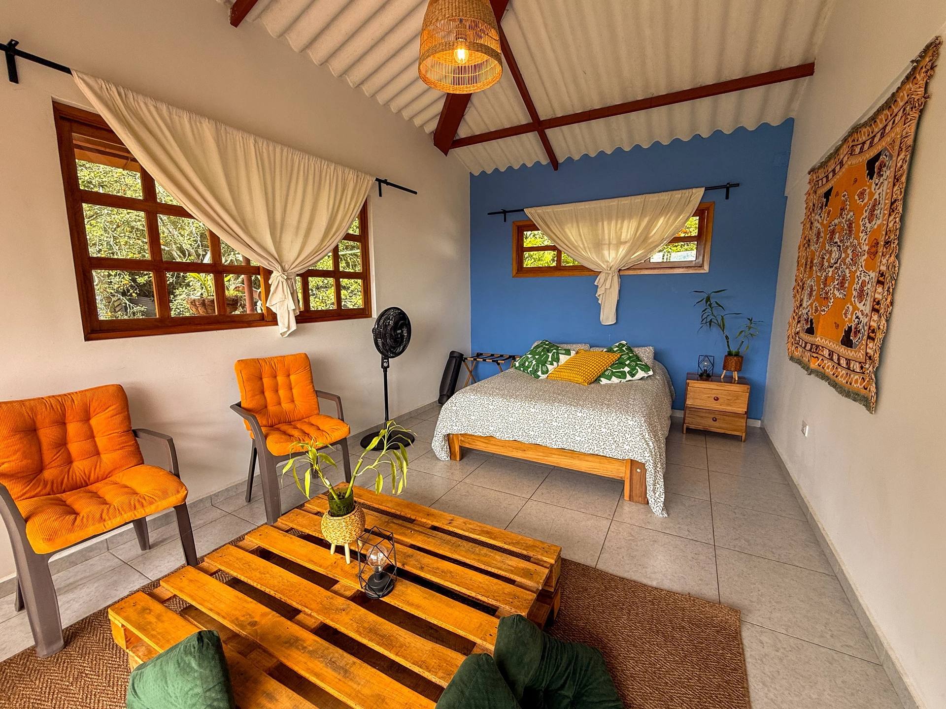 Interior de una habitación con ventanas de madera, cortinas blancas, paredes blancas y una pared azul, con una cama, sillas naranjas, una mesa de madera, una lámpara de café, plantas y una alfombra