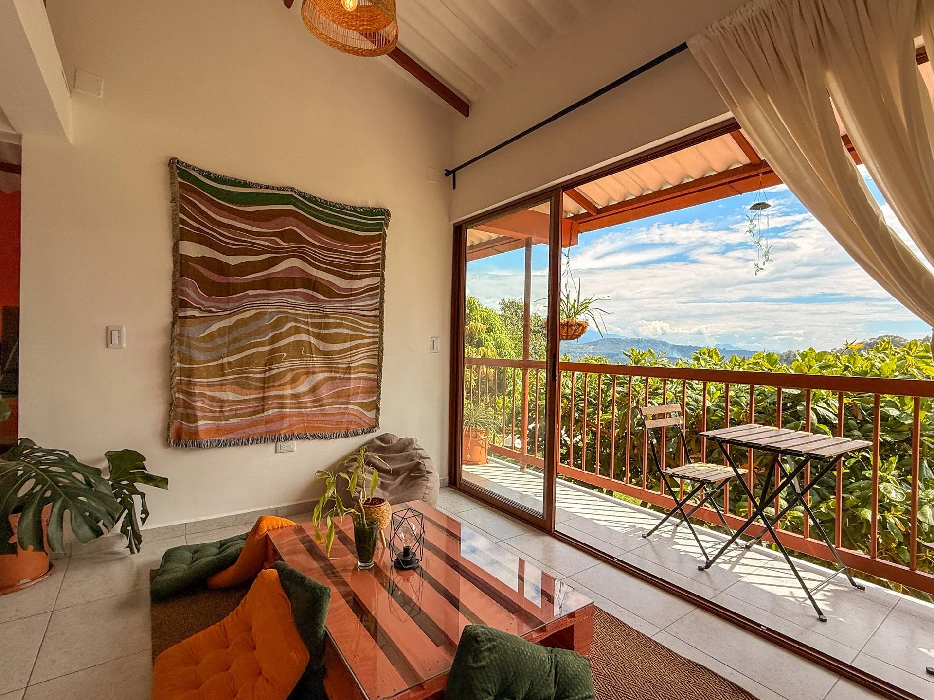 Sala de estar con ventanas grandes que dan a una terraza con vista a las montañas y cielo con nubes, decorada con un tapiz en la pared, plantas, cojines naranjas y verdes, mesa de vidrio y muebles de madera.