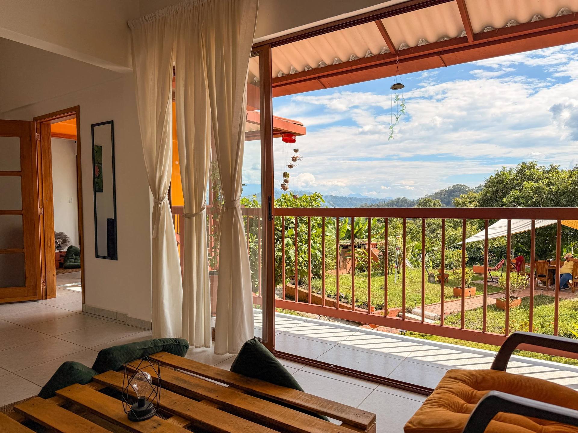 Interior de una casa con vista a un balcón y un paisaje natural bajo un cielo con nubes. Hay cortinas blancas, una mesa de madera y sillones de color verde y naranja.