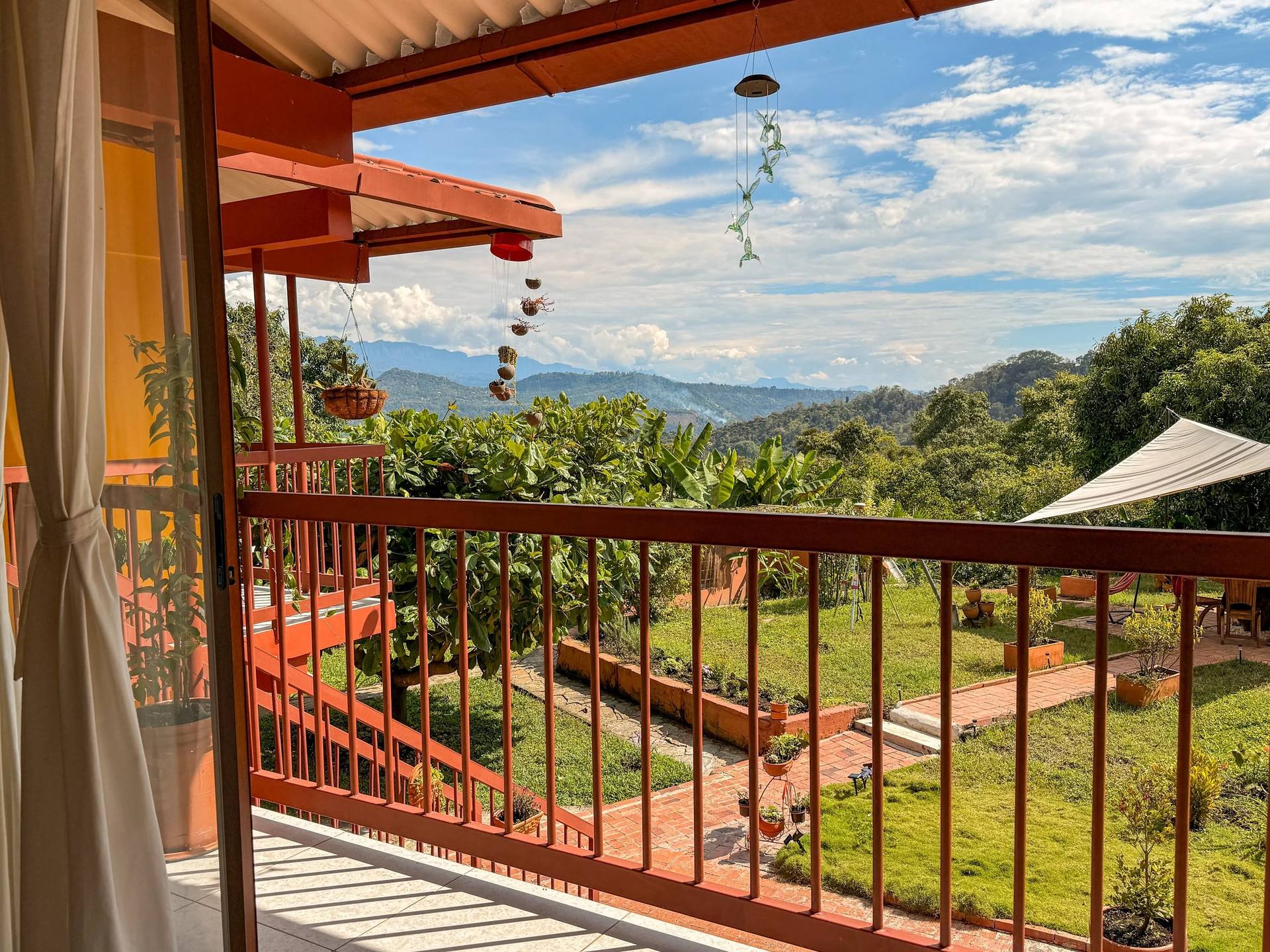 Vistas desde una terraza con un barandal de color rojo, que refleja un cielo azul con nubes blancas, rodeada de árboles verdes y montañas en el fondo.