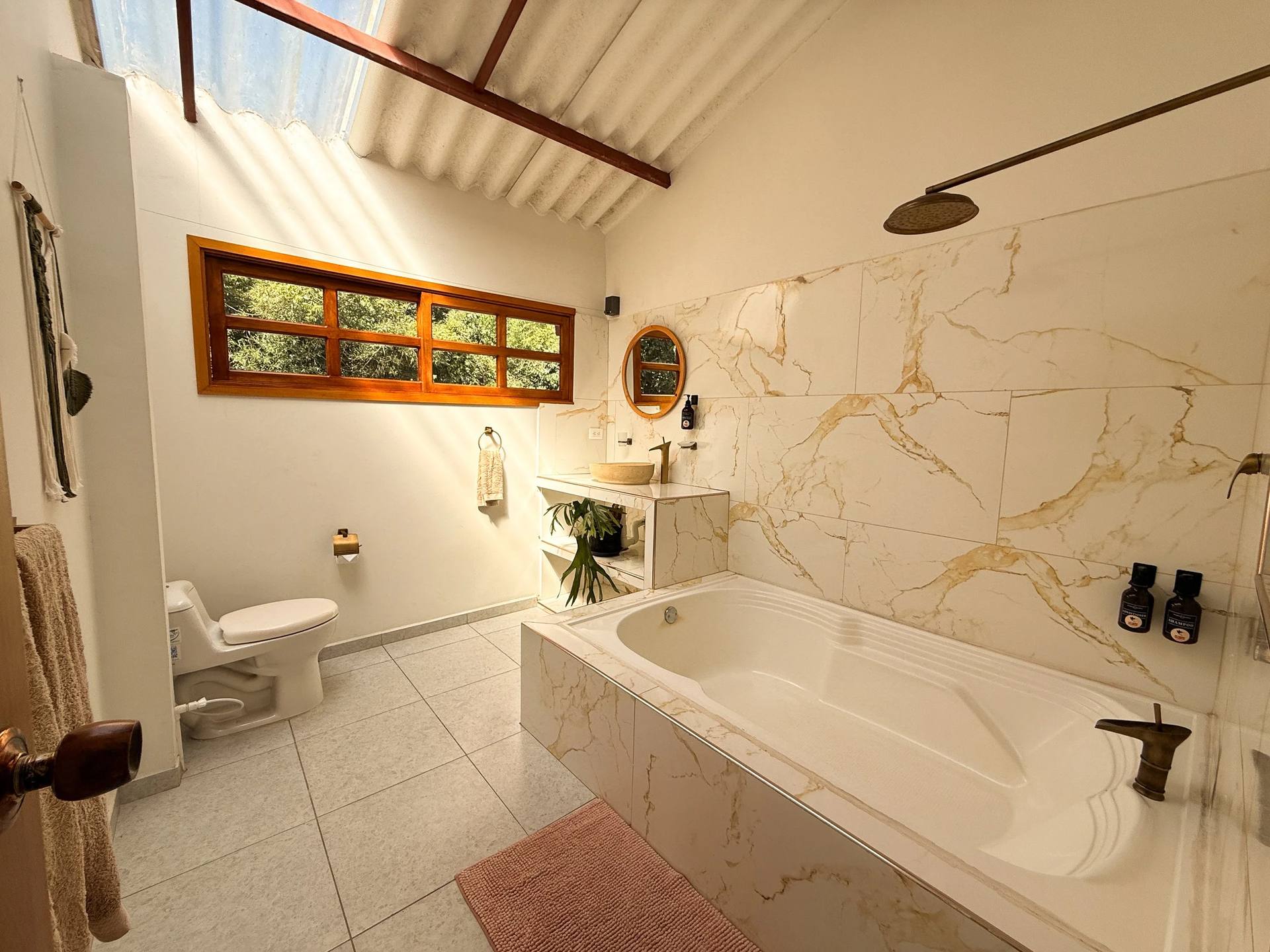 Un baño con una tina de baño de mármol, ducha de lluvia y ducha independiente, un inodoro, lavabo con espejo redondo y plantas decorativas, ventanas grandes con vista a la naturaleza, paredes blancas y piso de azulejo gris claro.