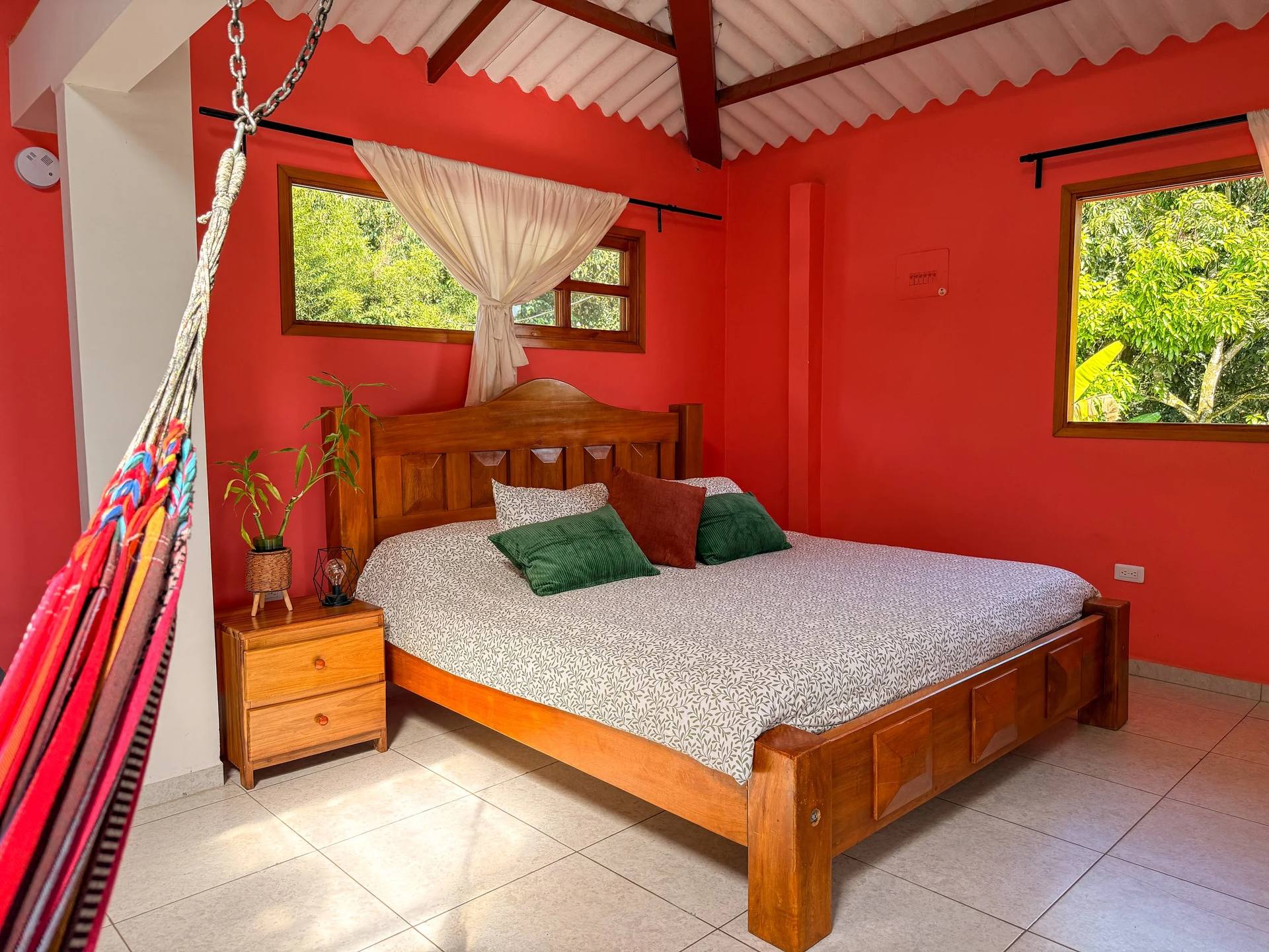 Vista de un dormitorio con paredes de color coral, una cama de madera con almohadas verdes y beige, una planta en una mesa de noche, ventanas con cortinas claras y un techo de tejas, en un entorno natural y luminoso.
