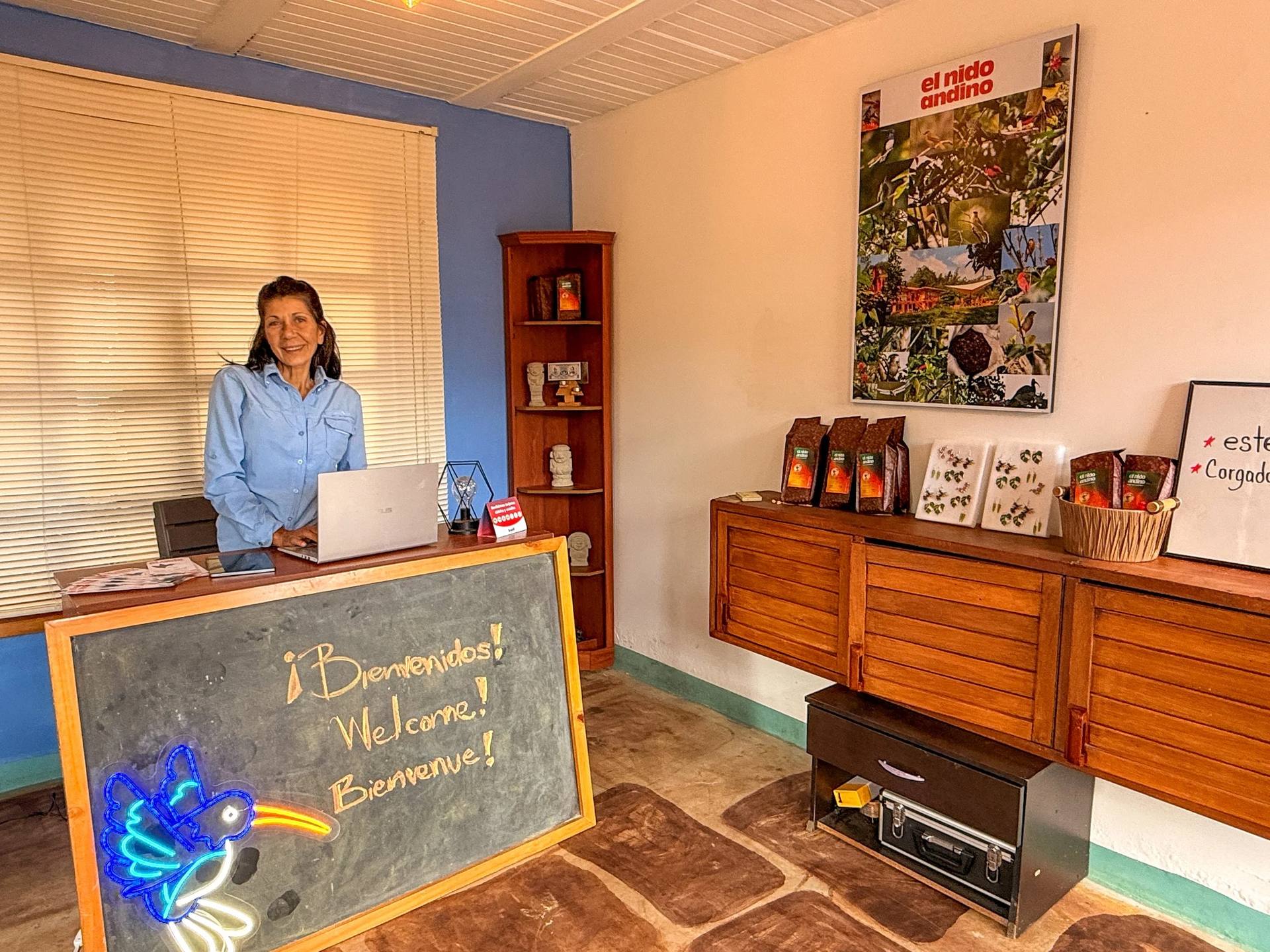 Una mujer sonriendo en un mostrador de una oficina con libro y señal de bienvenida en una pizarra. Decoración con plantas y un cartel grande de fotos sobre la cultura andina.