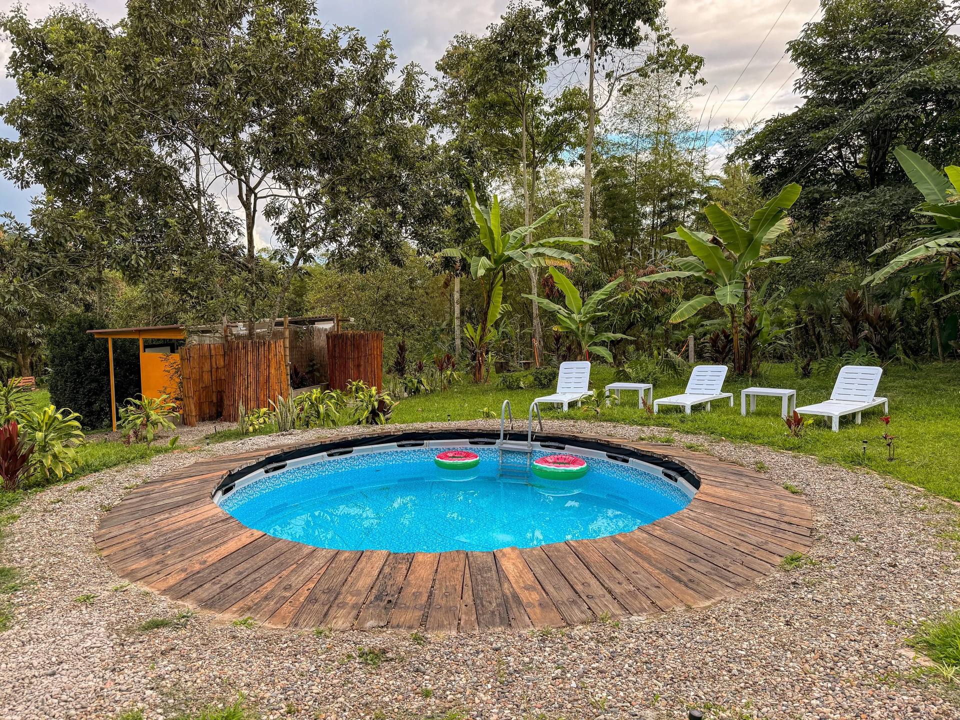 Piscina inflable en un jardín con sillas blancas, rodeada de árboles y plantas verdes, en un día nublado.
