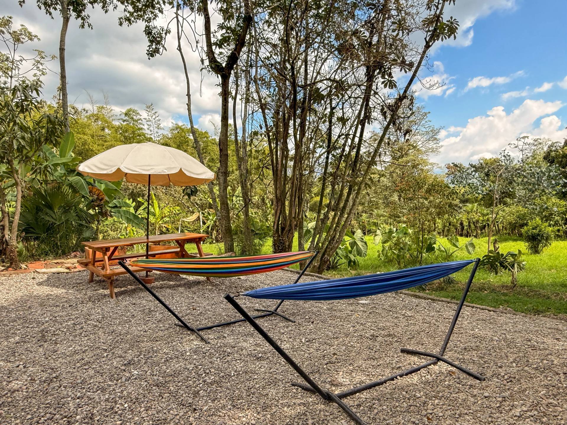 Un área exterior de descanso con hamaca, sombrilla, mesas de madera, sillas y muchas plantas y árboles verdes bajo un cielo parcialmente nublado.