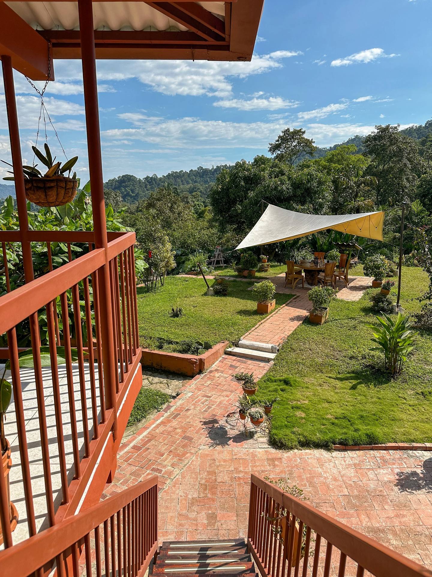 Vista desde una terraza roja con barandales, mostrando un jardín con árboles, plantas y mesas tapadas con una sombra, en un día soleado con cielo azul y algunas nubes.