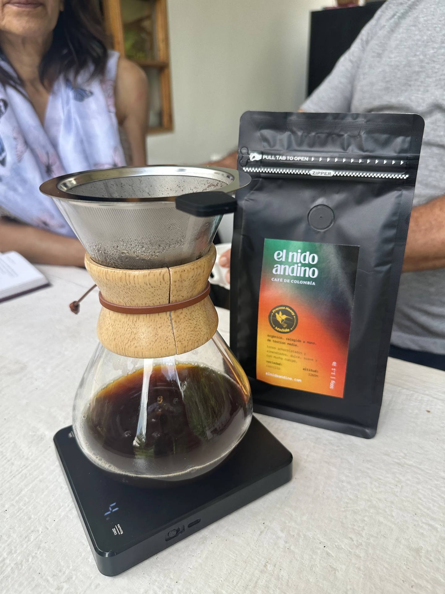 Telescopio de café con una cafetera de vidrio debajo, un paquete de café El Nido Andino y dos personas al fondo, una de ellas con un vestido blanco y la otra con una camiseta gris.