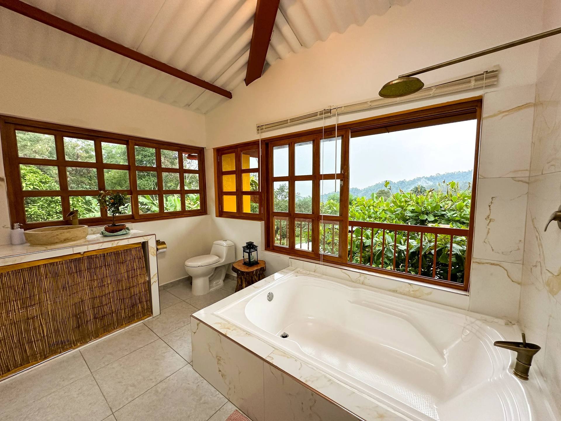 Baño con grandes ventanas de madera, vista a la naturaleza, bañera, lavabo de piedra, inodoro y decoración minimalista.