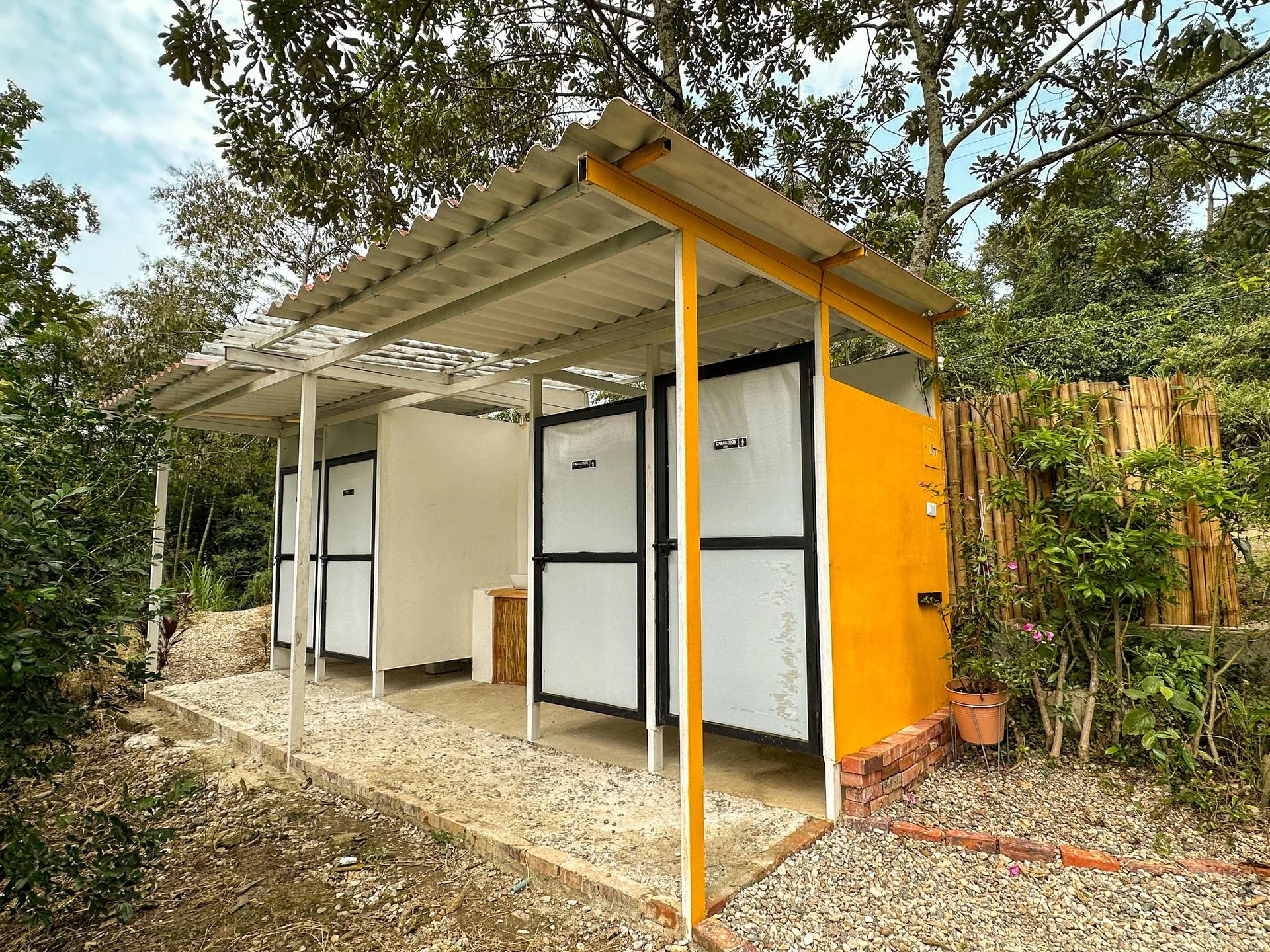 Vista de un baño público con puertas blancas y un marco de metal negro, una pared amarilla y un área cubierta con una estructura de techos de lámina metálica, rodeado de plantas y árboles.