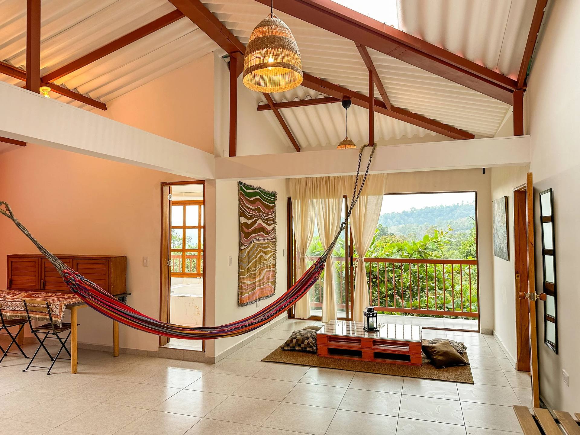 Interior de una sala con ventanales grandes, cortinas claras y decoración rústica, incluyendo hamaca, cuadros en la pared y un mueble de madera.