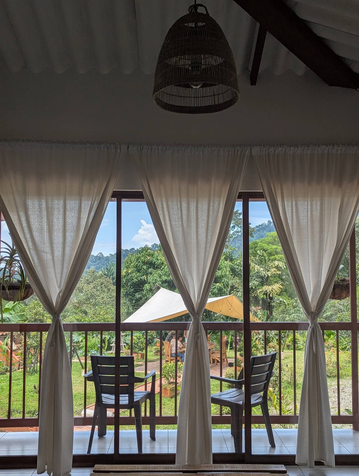 Vista desde una ventana con cortinas blancas, mostrando un patio con sillas y mesa, rodeado de vegetación y montañas al fondo bajo cielo azul.