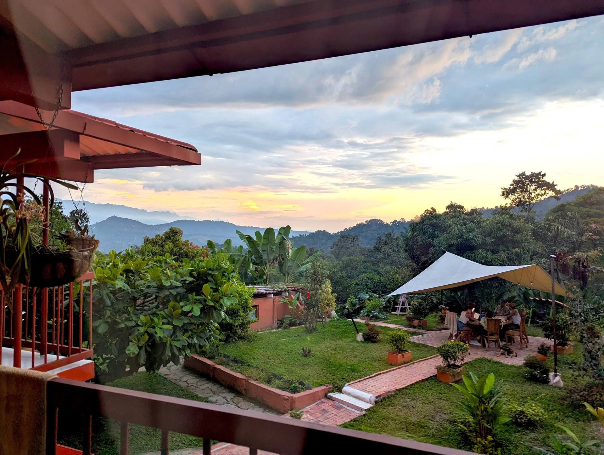 Vista de un jardín al atardecer con mesas al aire libre, árboles y montañas en el fondo, y un cielo con nubes.