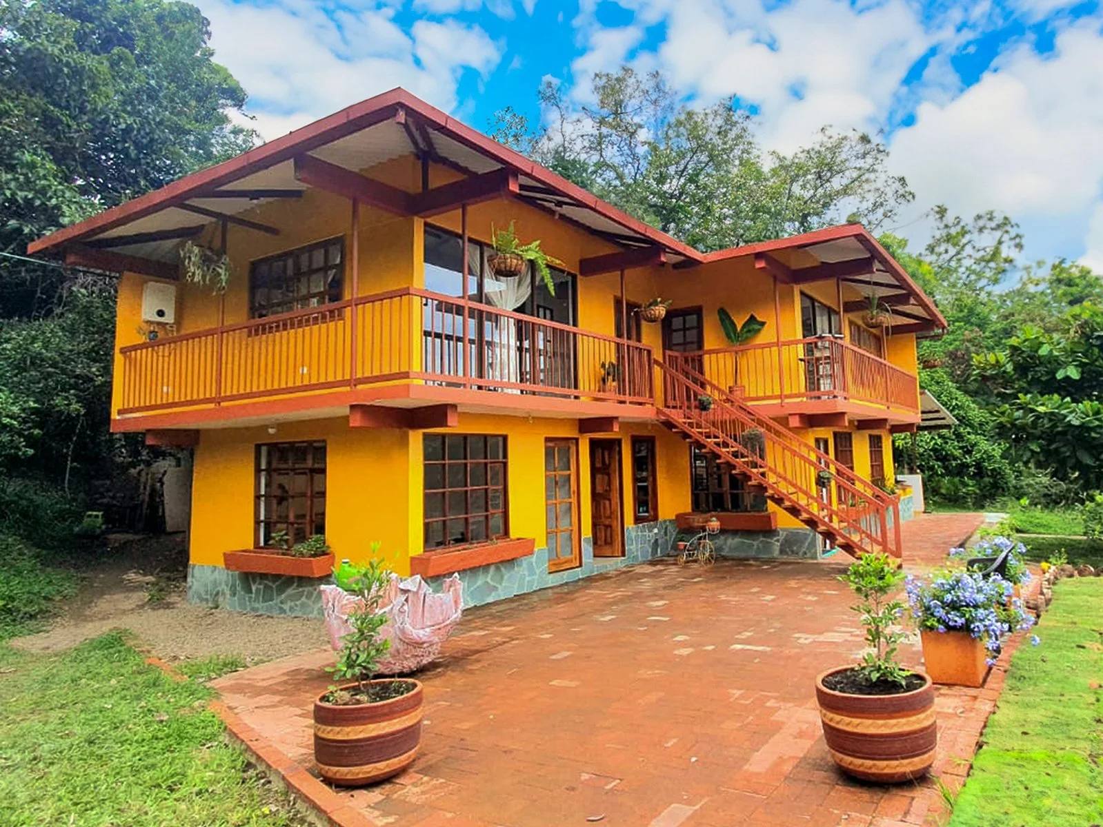 Casa color amarillo con cornisas y barandal naranjas, acceso externo con escaleras, rodeada de plantas y macetas en un entorno natural con árboles y cielo azul.